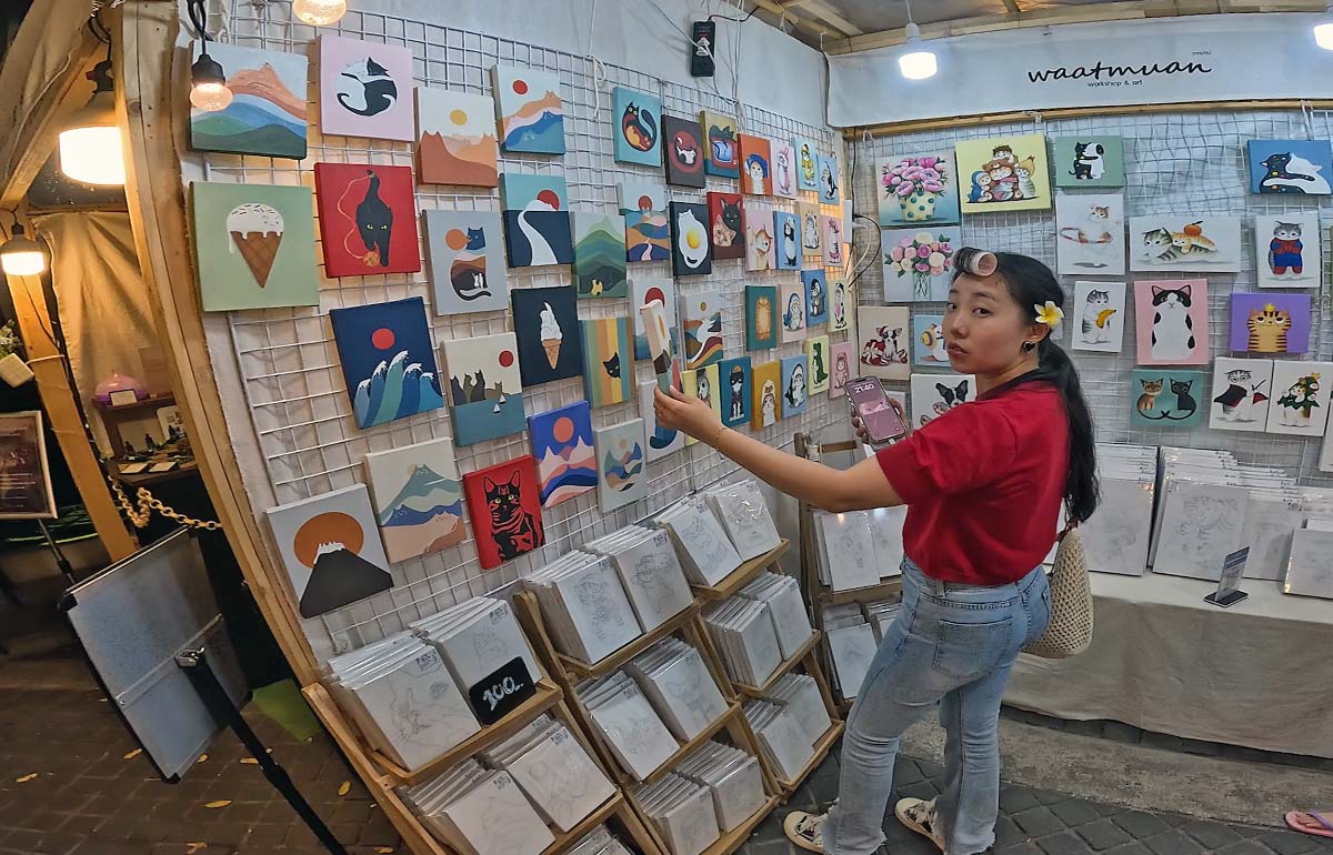 Young woman with a curler in her hair observing a painting being sold at the White Market, one of Nimman night markets in Chiang Mai, Thailand; photo by Ivan Kralj.