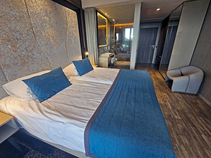 Guestroom interior in Hotel Jama, Postojna, Slovenia, with a large bed with blue pillows and bed runner, the rest of the room in beige and gray tones. En suite bathroom is behind the see-through glass; photo by Ivan Kralj.