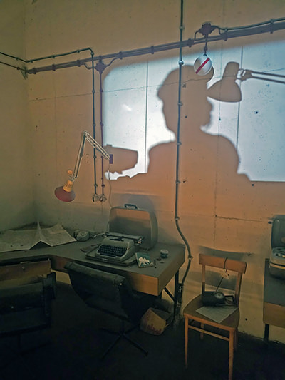 A shadow of a typist projected on the wall behind typewriter desk displayed in one of the Secret Rooms, a former surveillance center at Hotel Jama, in Postojna, Slovenia; photo by Ivan Kralj.