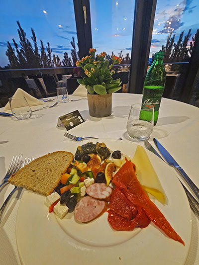 Appetizer containing some Carniolan sausage, cheese, and salad, with Radenska mineral water on the side, served on a table of Magical Garden Restaurant in Hotel Jama, Postojna, Slovenia, during sunset; photo by Ivan Kralj.