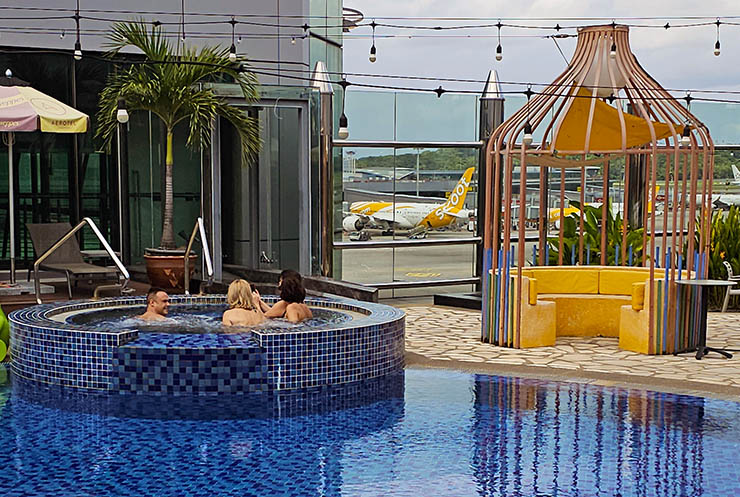 Passengers resting in a jacuzzi by the pool, on the roof of Singapore Chanig Airport's Terminal 1 building, with a plane parked in the background; photo by Ivan Kralj.