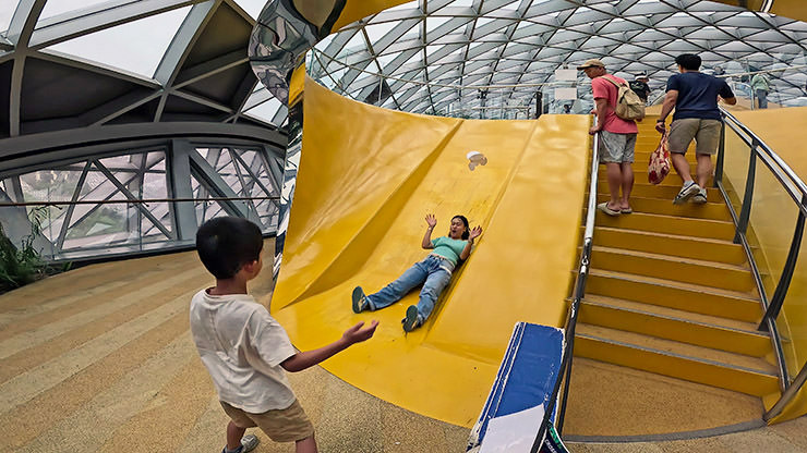 An adult woman sliding down the yellow Discovery Slide with surprise on her face, at Canopy Park, Jewel Changi Airport in Singapore; photo by Ivan Kralj.